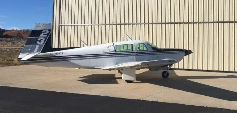 A white and black Grumman American AA-5 Traveler aircraft, N1047A, parked on concrete next to a hangar.