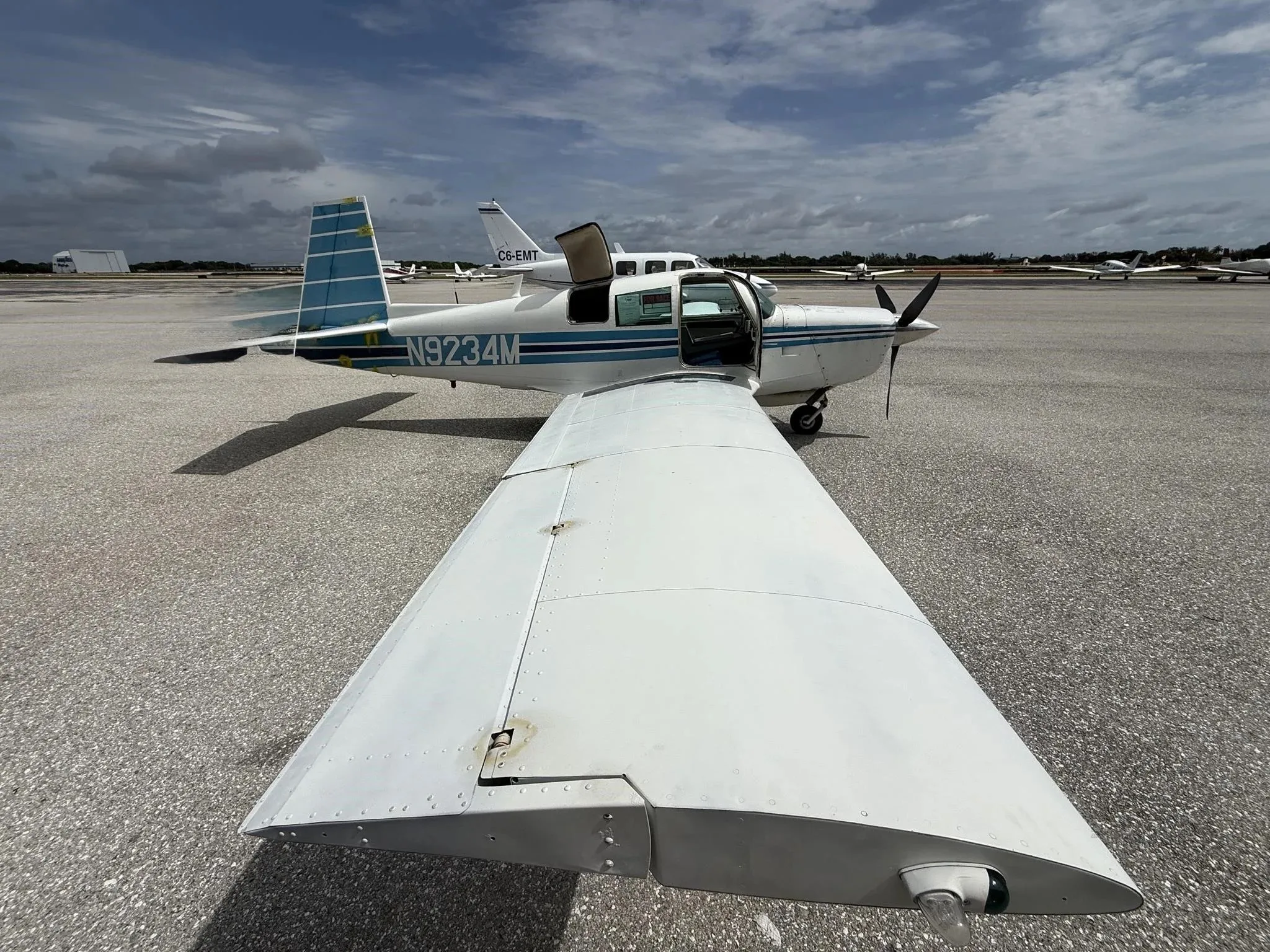 A white single-engine aircraft with blue stripes, registration N9234M, parked on an airfield with its door open under a partly cloudy sky.