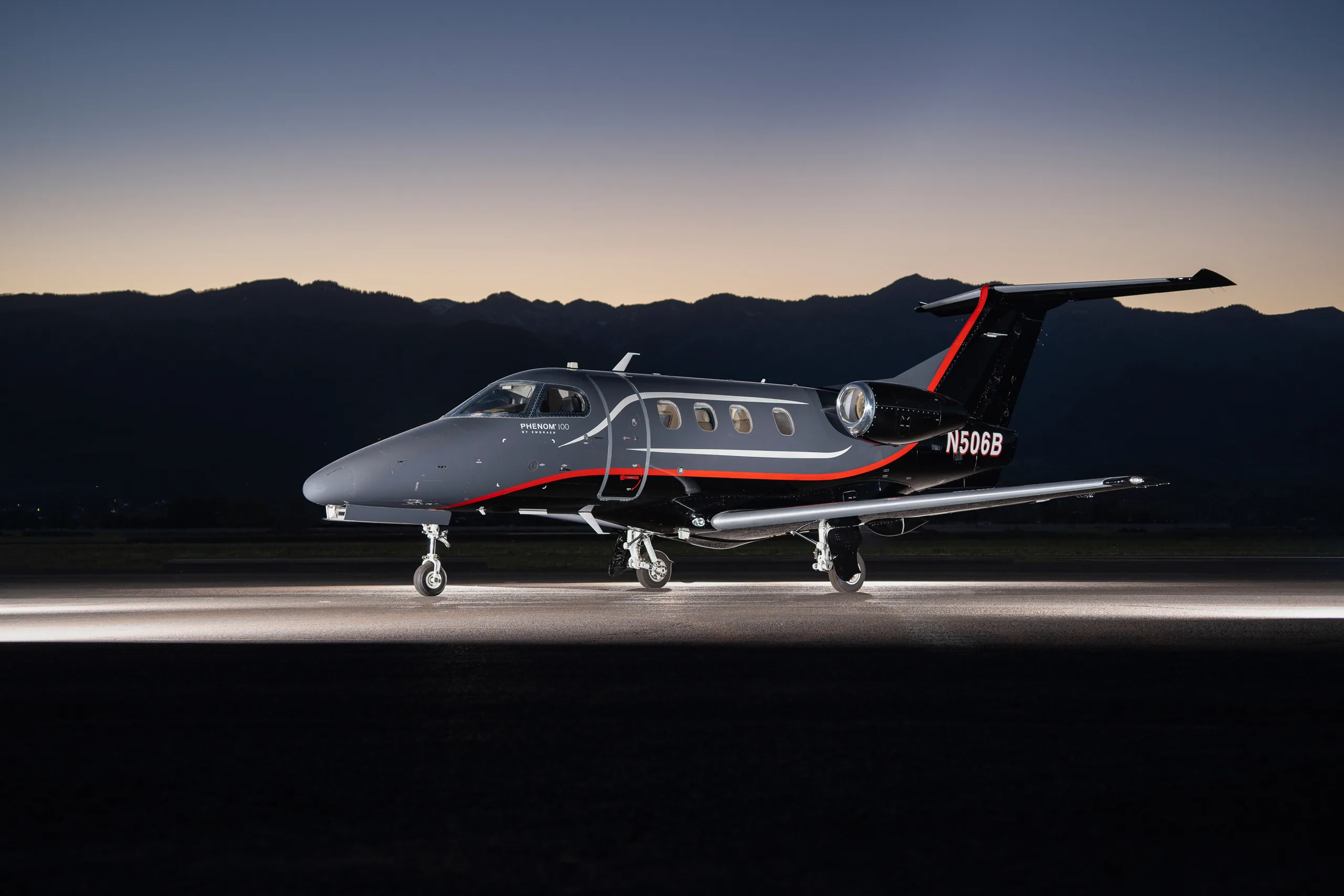 A gray and black Embraer Phenom 100 business jet with red and orange stripes, registration N506B, parked on a tarmac at dusk with mountains in the background.