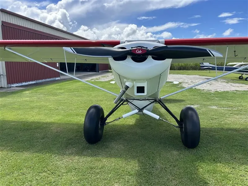 A head-on view of the white and red Super Cub aircraft with its propeller and large tundra tires, parked in front of a hangar.