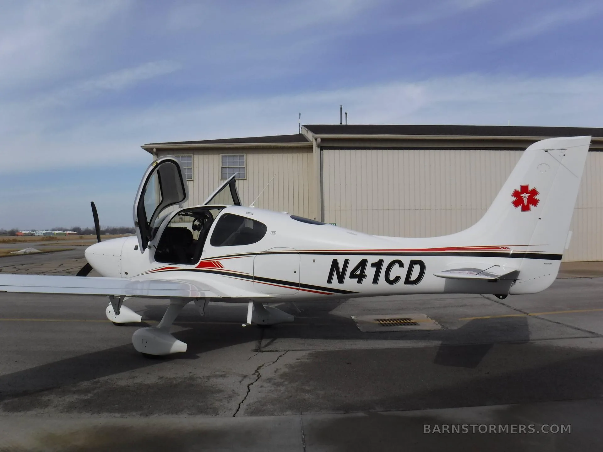 A white Cirrus SR22 aircraft with red and black stripes and an EMS star on the tail, parked on an asphalt ramp with its doors open.