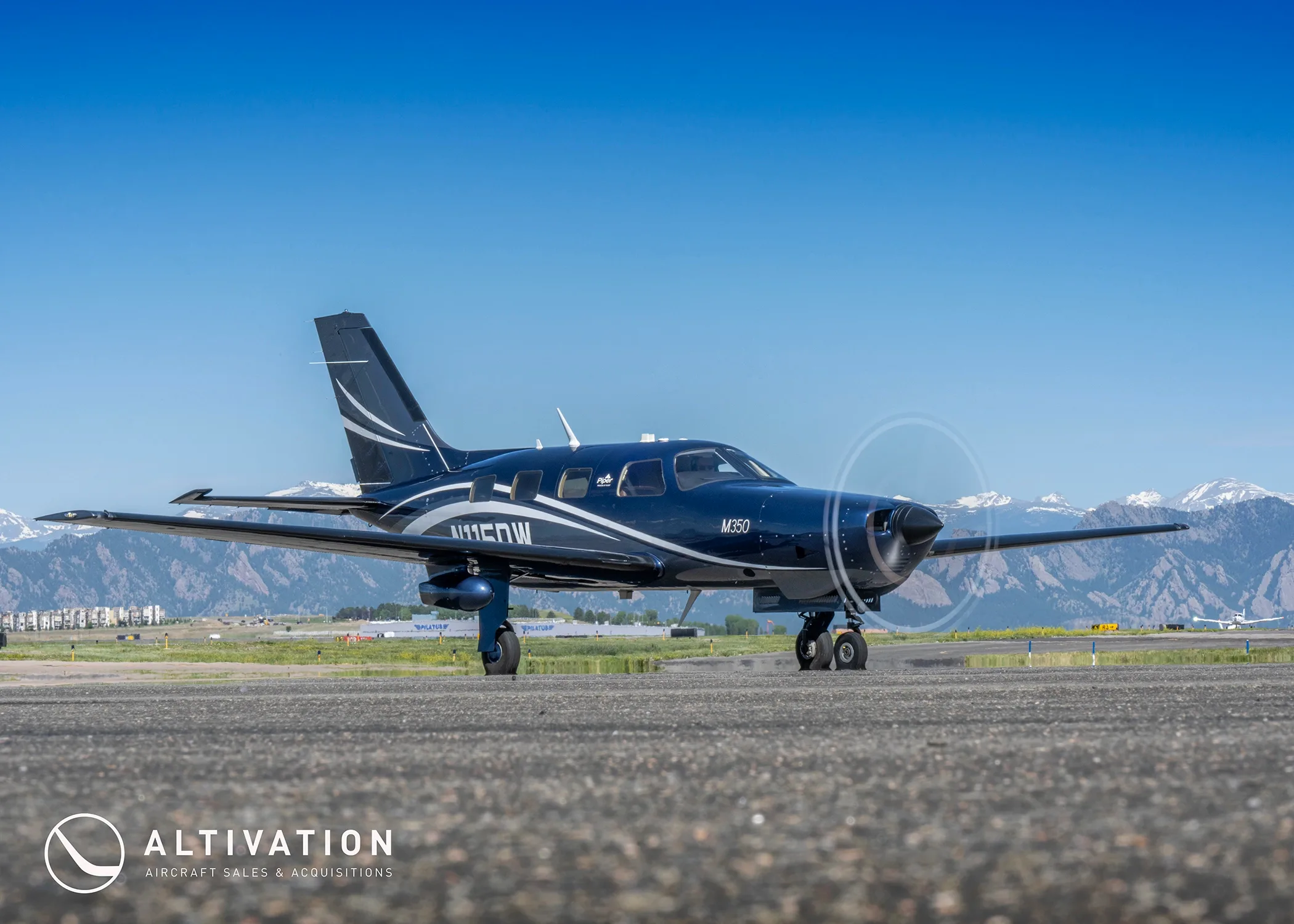 A dark blue Piper M350, tail number N115DW, on a tarmac with mountains and other aircraft in the distance.