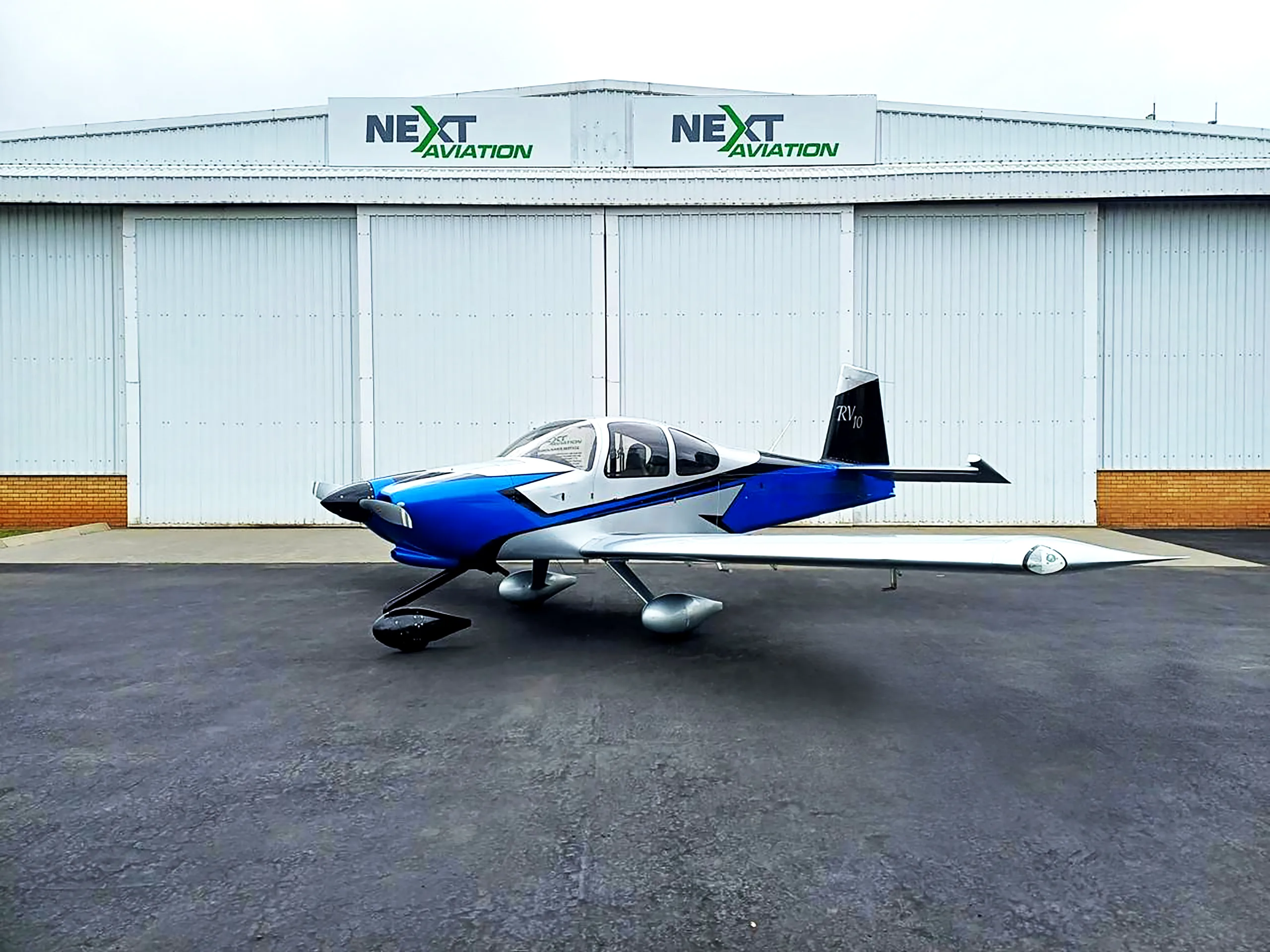 A blue and silver RV10 aircraft is parked on the tarmac in front of a white hangar with "NEXT AVIATION" signs.