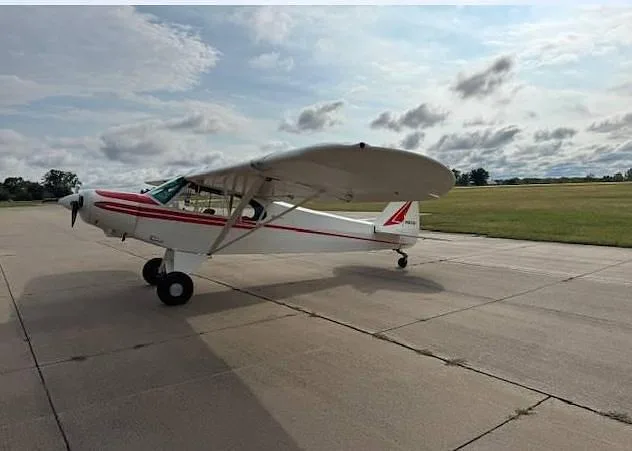 A white and red single-engine taildragger aircraft is parked on a concrete tarmac next to a grassy field.