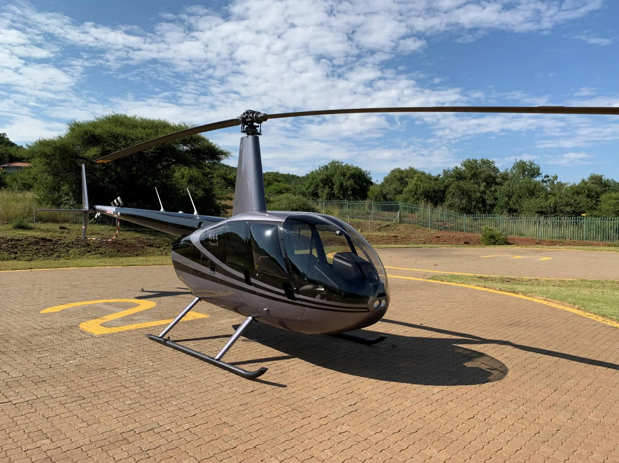 A grey and black Robinson R44 Raven II helicopter is parked on a paved helipad with a yellow '2' marking, under a partly cloudy sky.