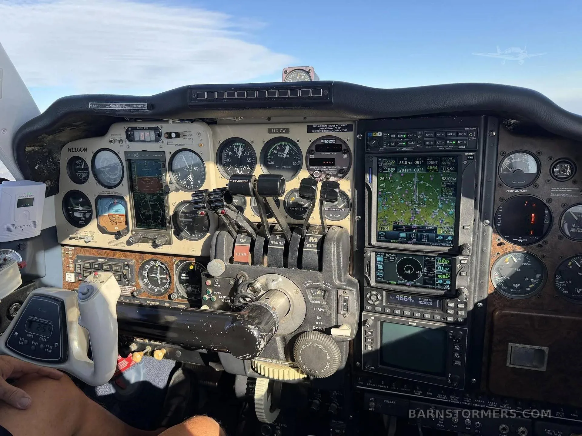 The cockpit of an aircraft showing the instrument panel with analog gauges, digital displays, and flight controls, with a person's leg visible.