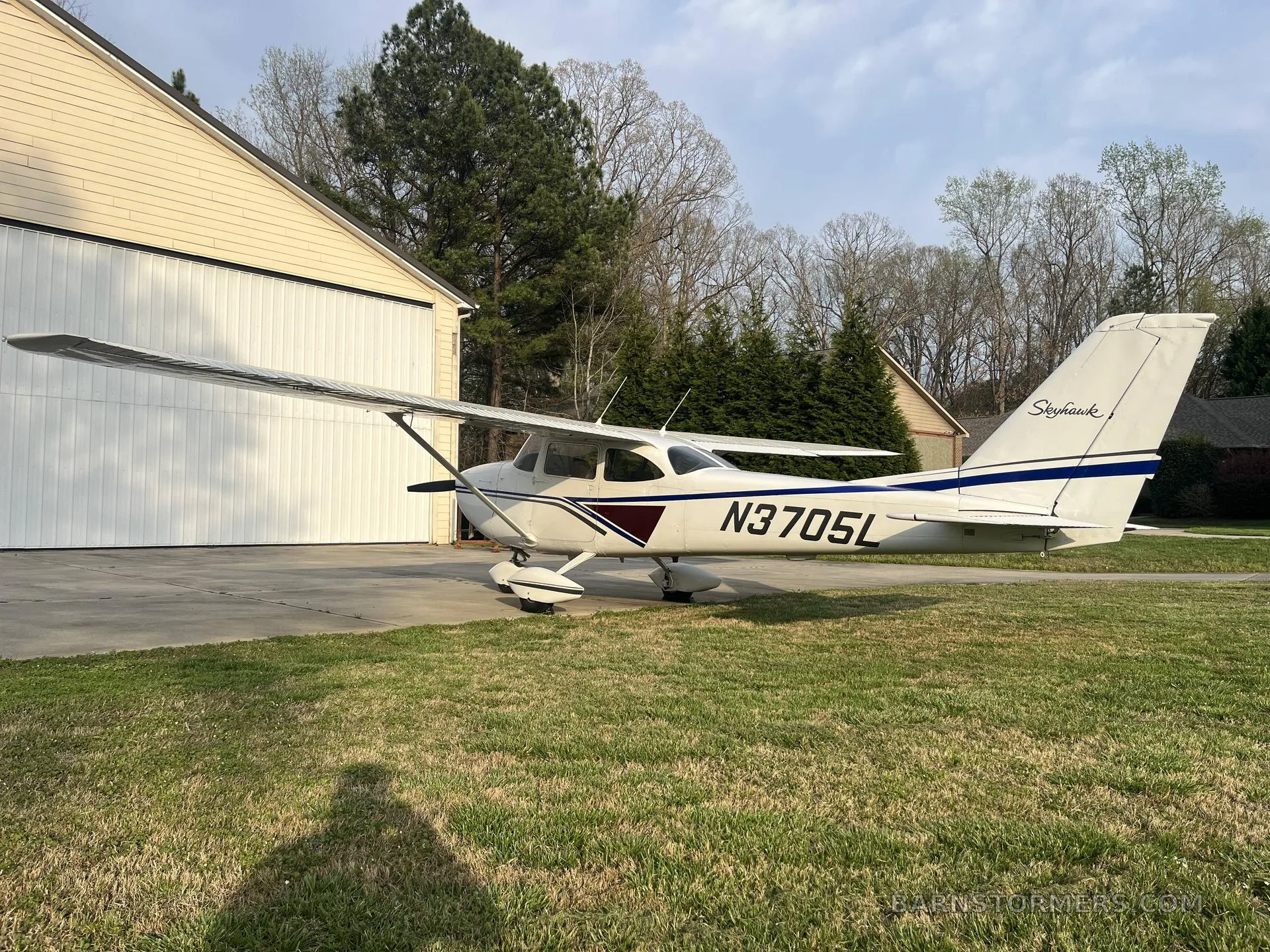A white Cessna Skyhawk (N3705L) with blue and maroon stripes is parked on grass in front of a white hangar.