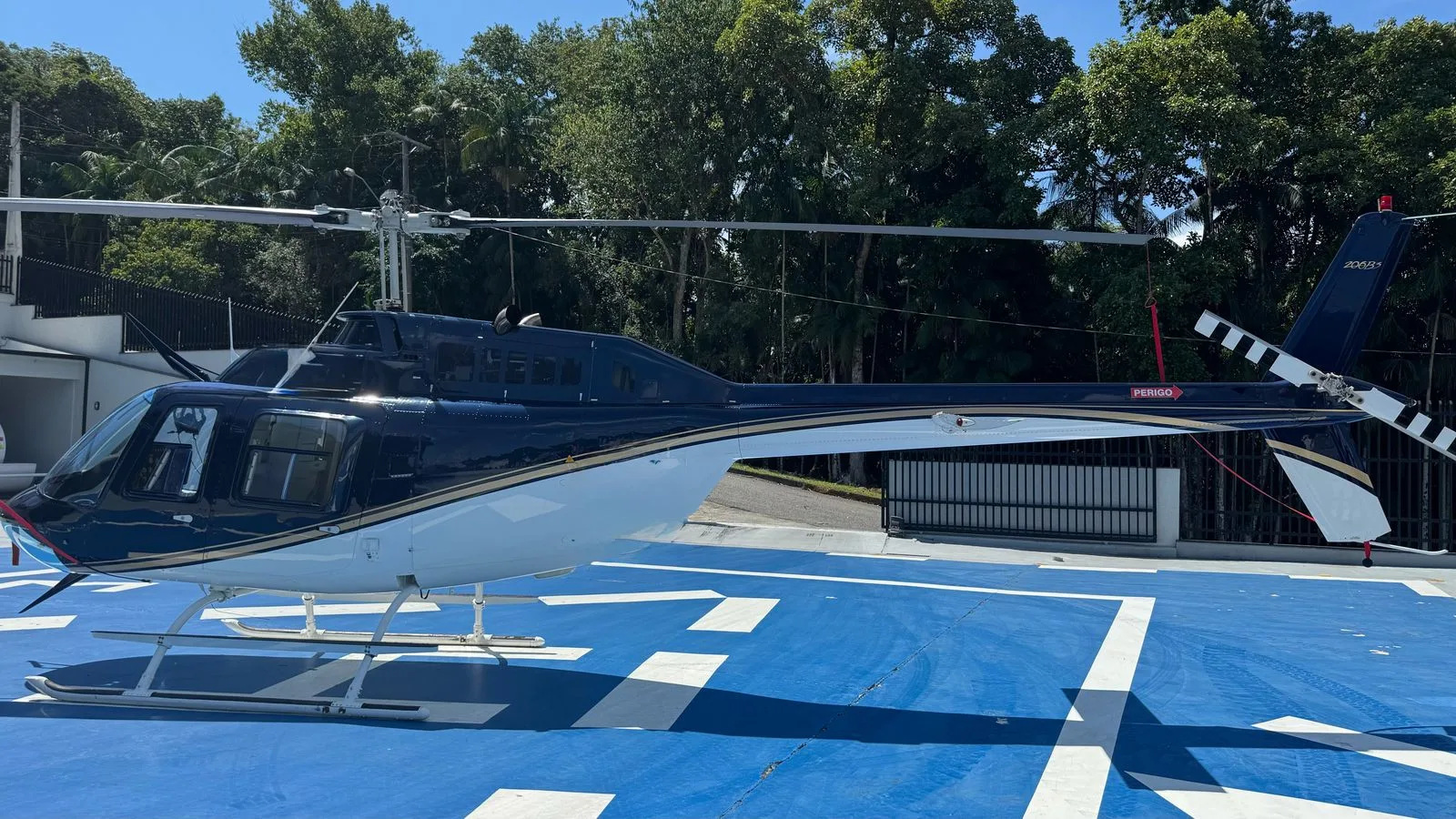 A side view of a dark blue and white helicopter with '206B3' on the tail, parked on a blue helipad.