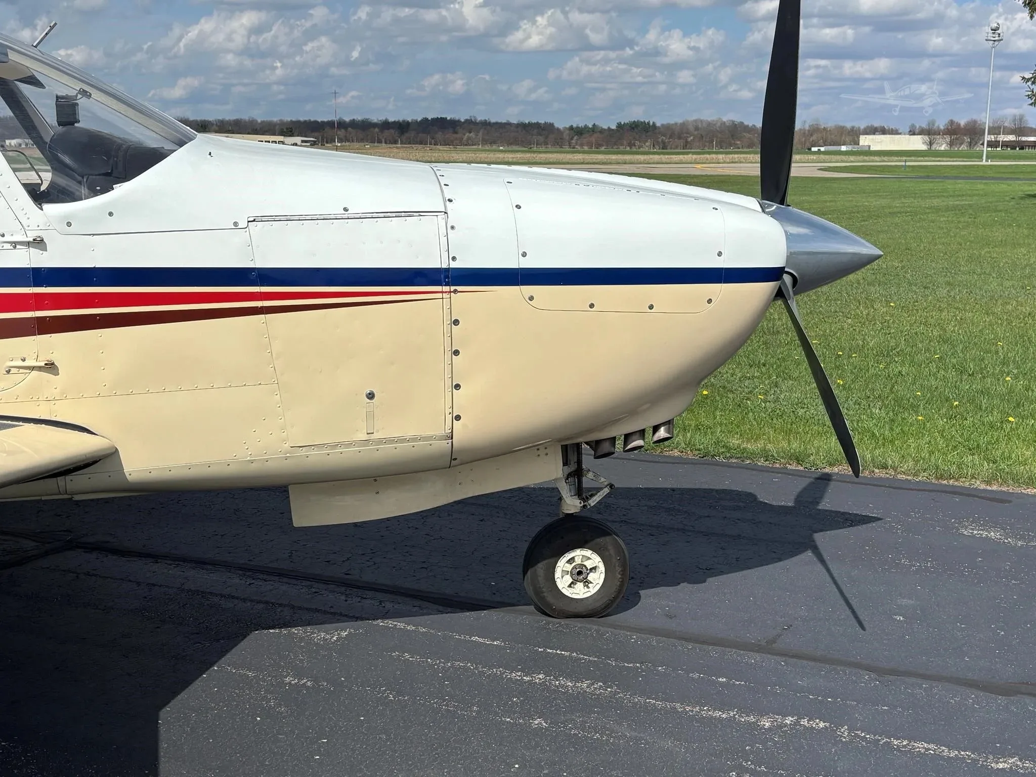A close-up side view of the nose and front landing gear of a white and beige single-engine aircraft.