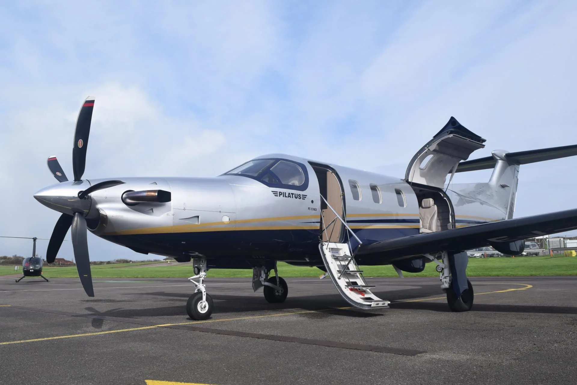A silver Pilatus PC-12 NGX with blue and gold stripes sits on an airfield tarmac with its doors open, a helicopter visible in the background.