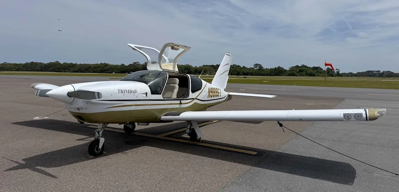 A front-quarter view of the white and gold Socata TB-20 Trinidad aircraft with its doors open on an airfield.
