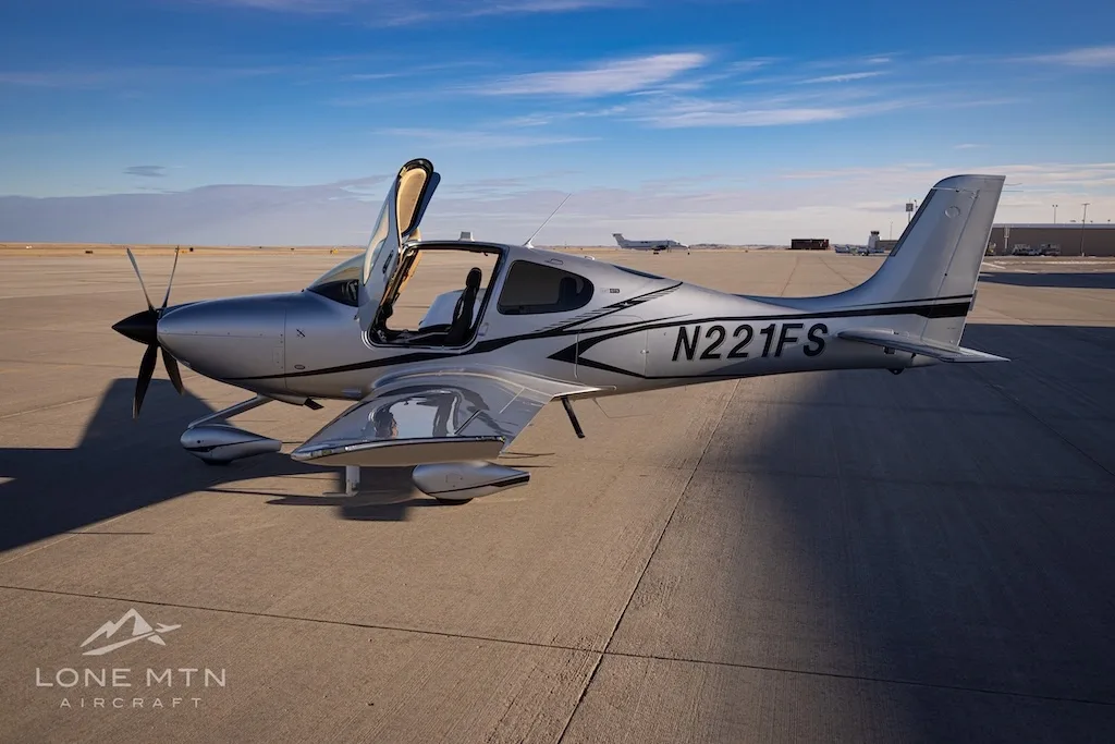 A side view of the silver Cirrus SR22T aircraft N221FS with its doors open, parked on an airfield with another plane in the distance.