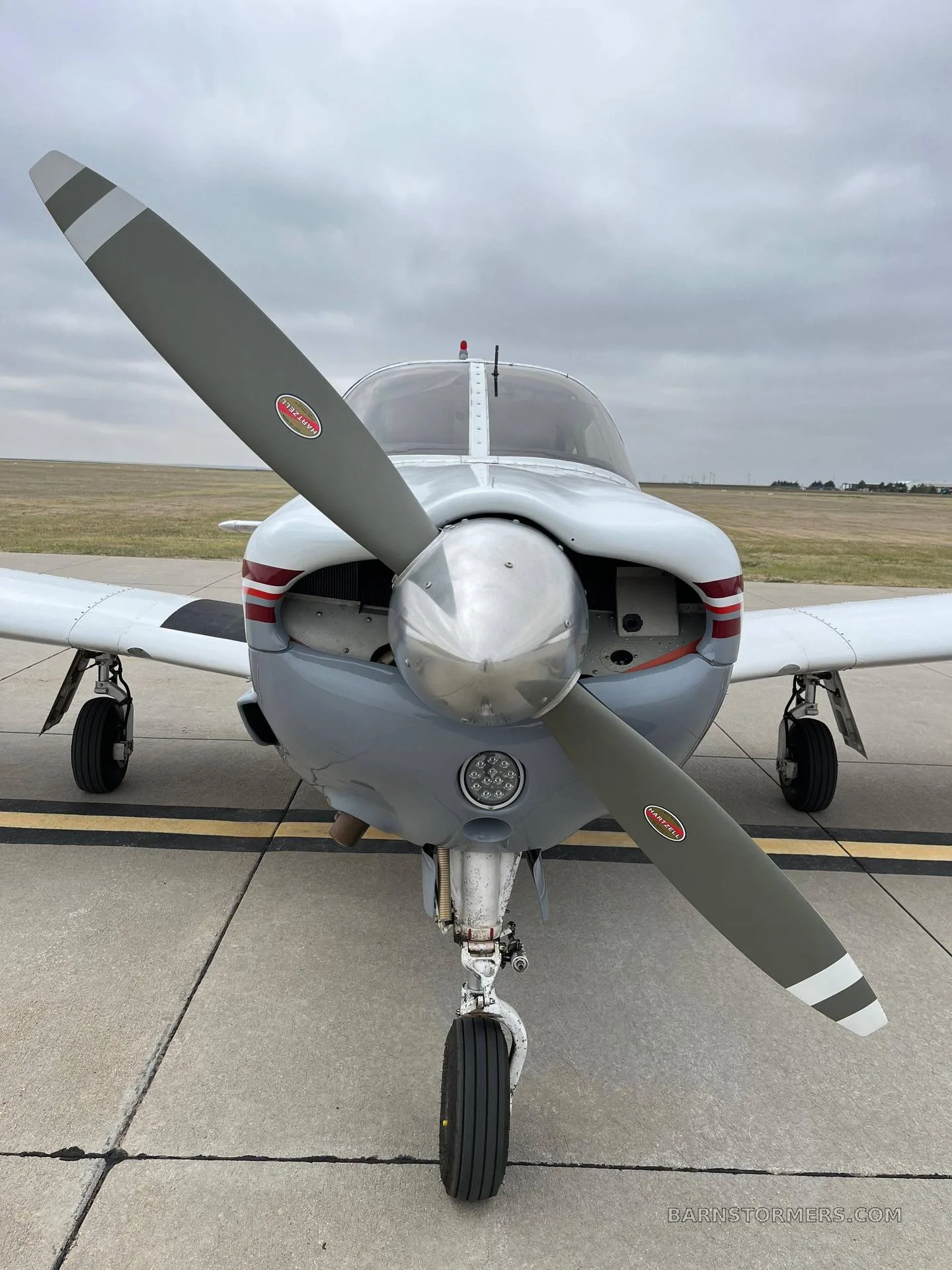 Front view of a light gray and white single-engine aircraft with a red stripe, showing the propeller and nose gear, parked on an asphalt ramp.