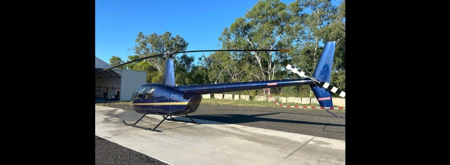 A dark blue Robinson R44 helicopter with a gold stripe parked on a concrete pad next to a hangar.