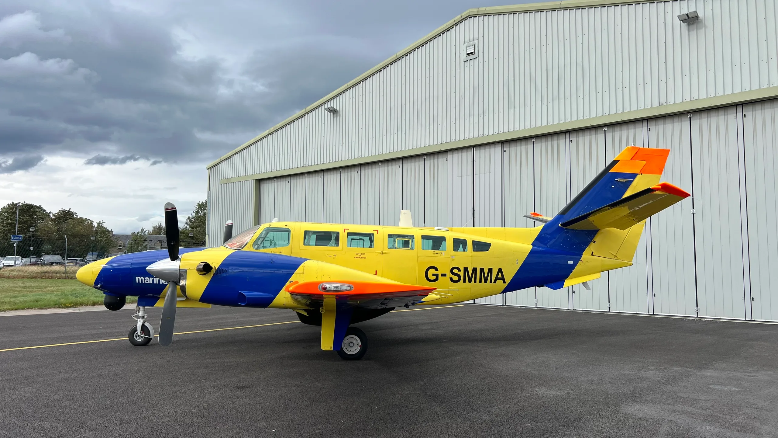 A yellow and blue twin-engine propeller aircraft with registration G-SMMA is parked on an airfield next to a white hangar under a cloudy sky.