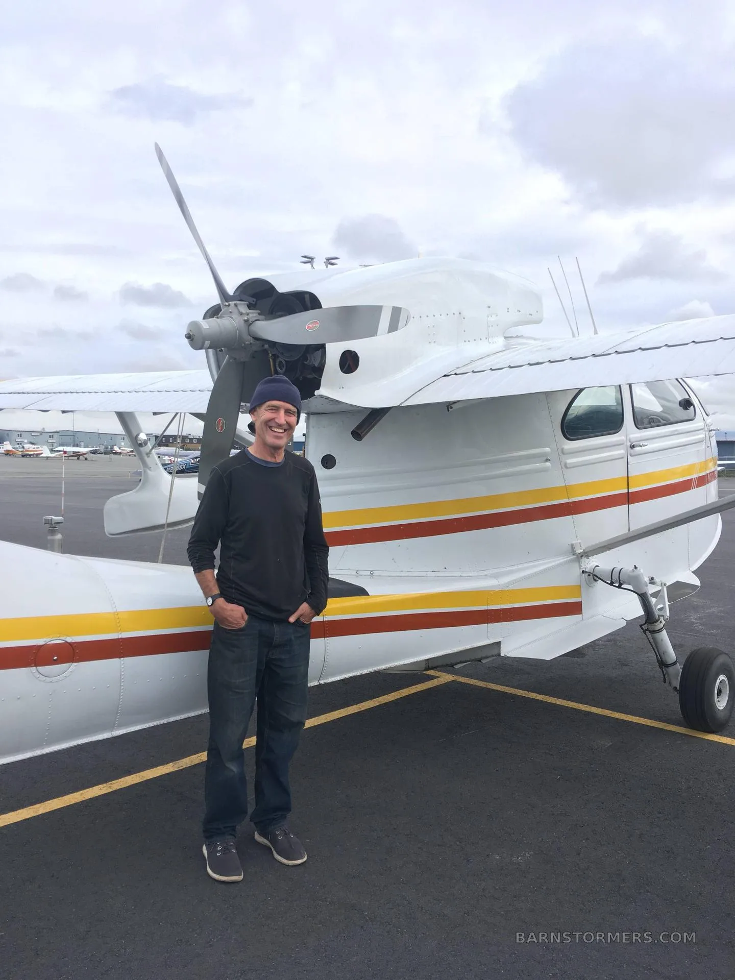A man in a dark shirt and jeans stands next to the white Republic RC-3 Seabee seaplane on an airport tarmac.