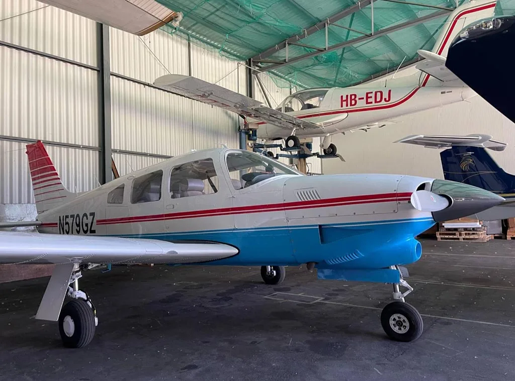 A white and blue Piper PA-28-181 Archer II with red stripes, parked inside a hangar with other aircraft.