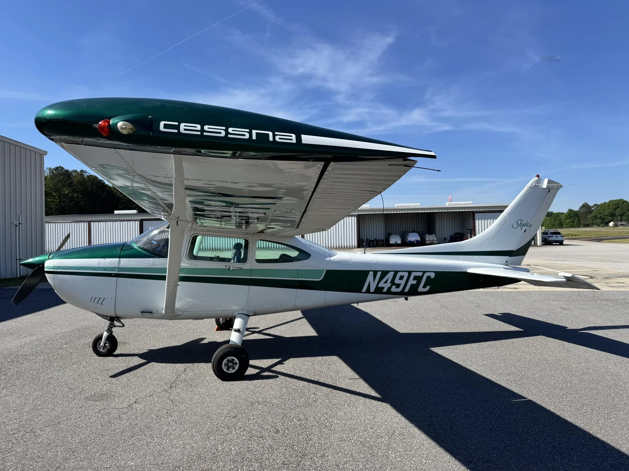 A side view of the white and green Cessna 182 Skylane, N49FC, parked on an airfield with hangars in the background.