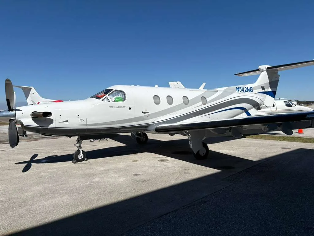 A white Pilatus PC-12 NG aircraft with blue and grey stripes, registration N542NG, parked on a tarmac under a clear blue sky.
