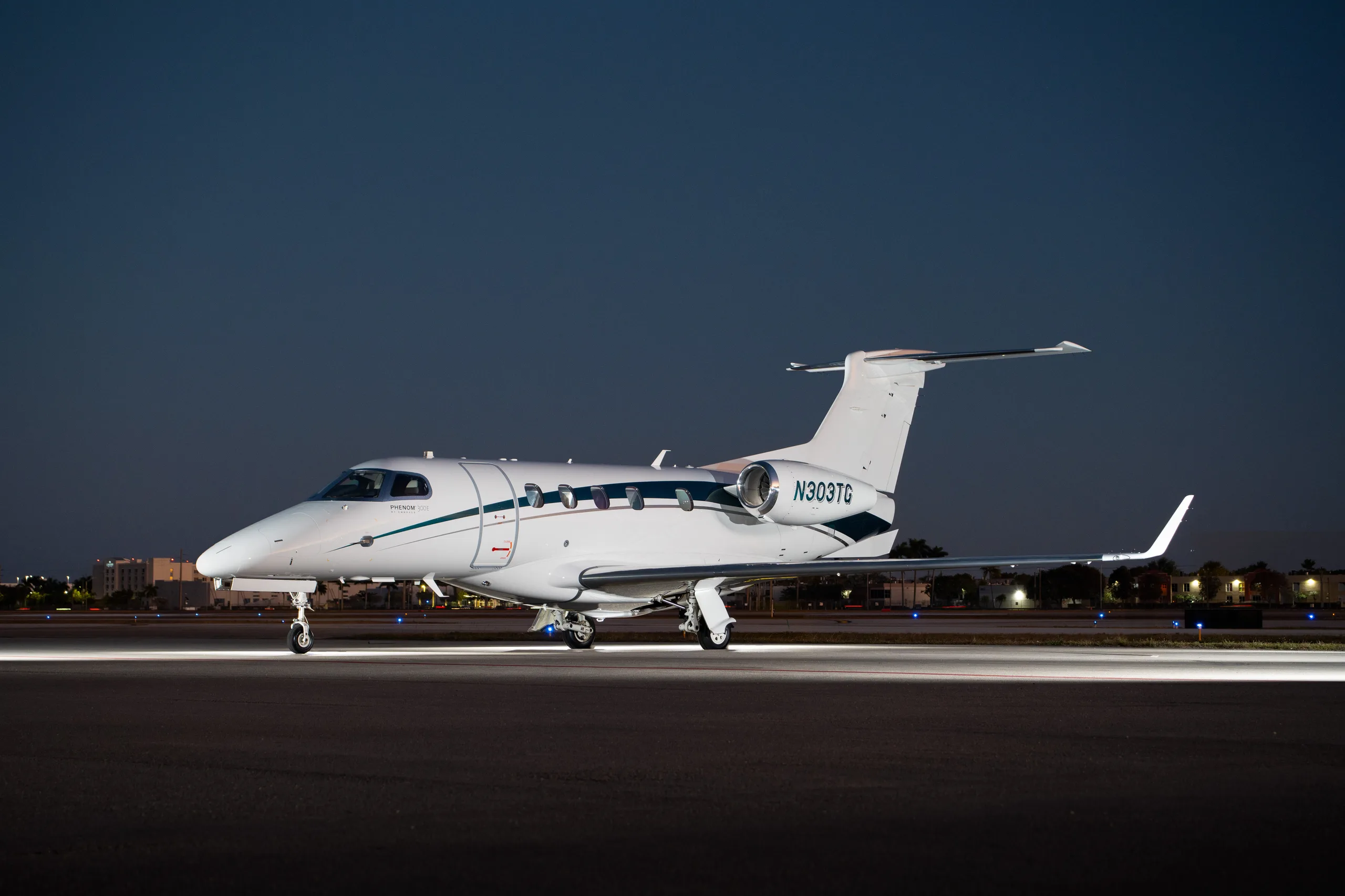 A white Embraer Phenom 300 business jet with dark blue and green stripes, registration N303TG, sits on a tarmac at dusk.