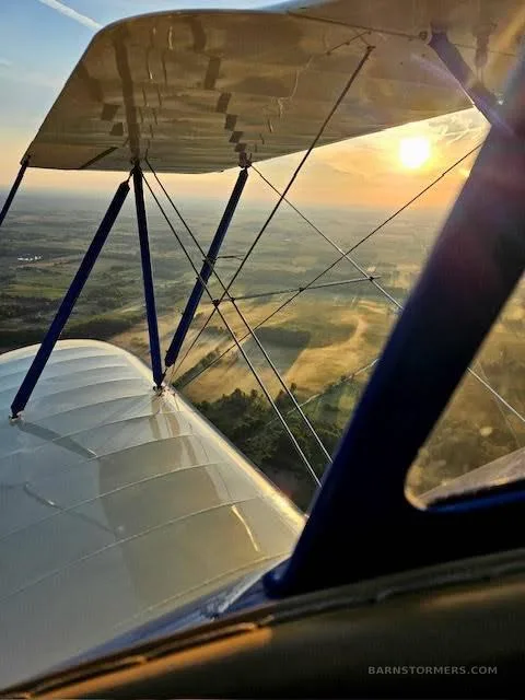 A view from a biplane's cockpit looking over the wing at a sunset landscape below.
