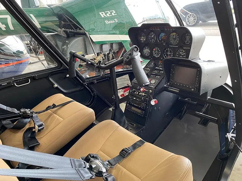 The cockpit of a Robinson R44 helicopter features tan seats, a cyclic stick, and an instrument panel with gauges and a GPS unit.