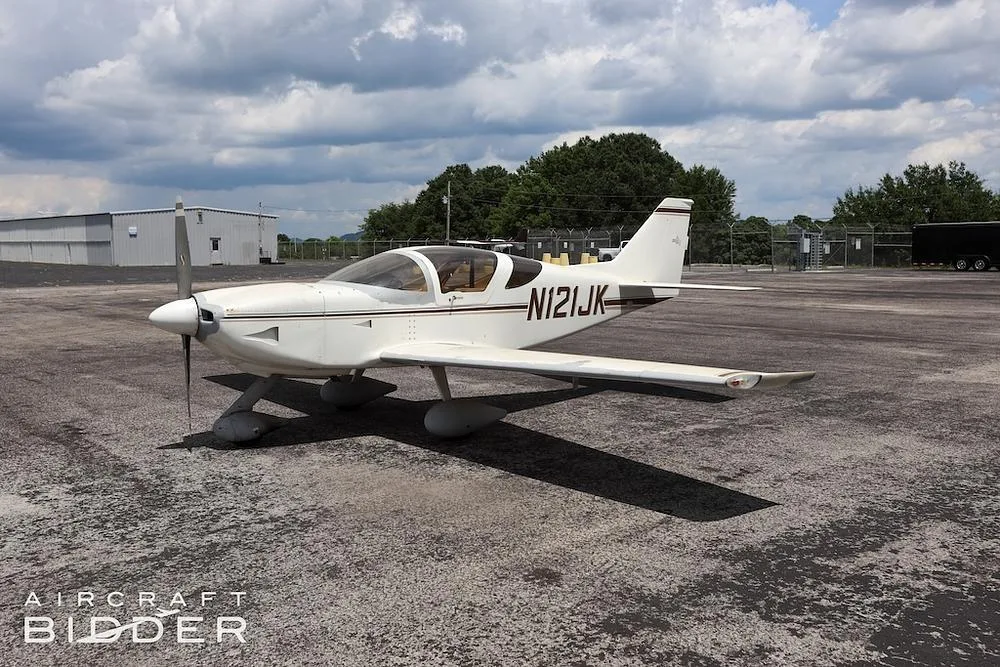 A white single-engine aircraft (N121JK) with brown stripes parked on an asphalt apron under a cloudy sky.