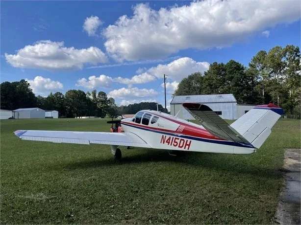 A white and red Beechcraft Bonanza N415DH parked on grass under a blue sky with scattered clouds.