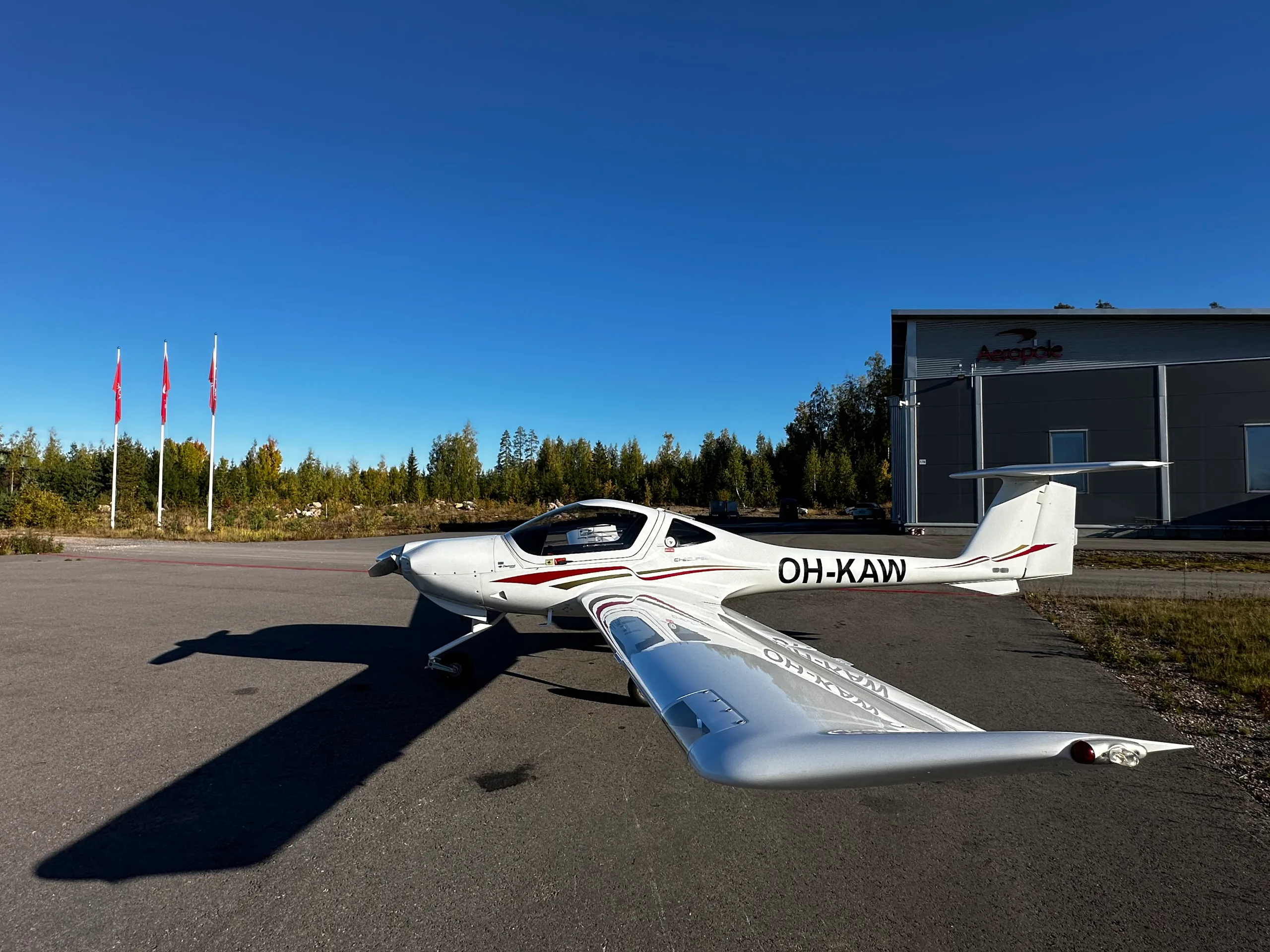 A white Diamond DA20 aircraft with registration OH-KAW and red/black stripes parked on an asphalt apron next to a hangar.