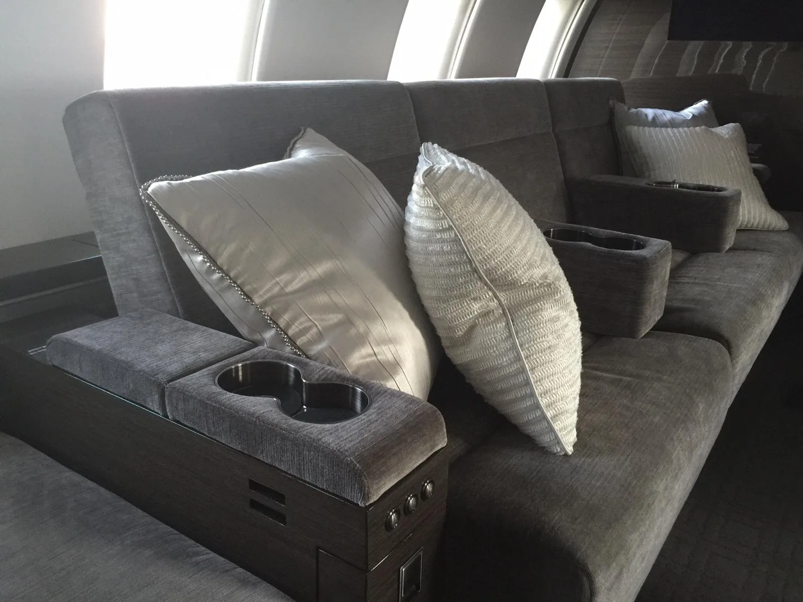 A comfortable grey sofa with white pillows and integrated cup holders inside a private jet cabin.
