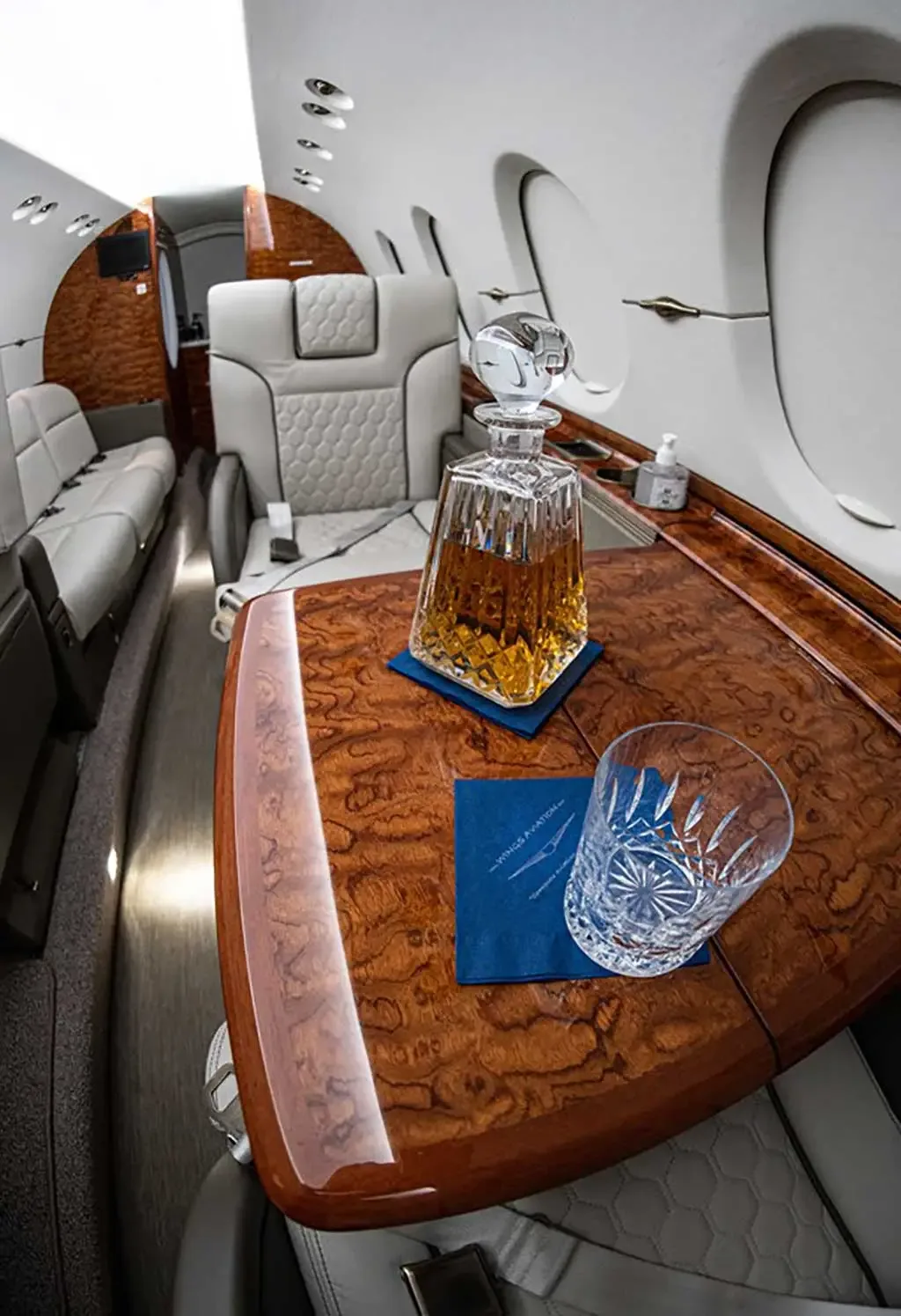 A close-up of a polished wood table in a private jet cabin, featuring a crystal decanter and glass, with light grey seats in the background.