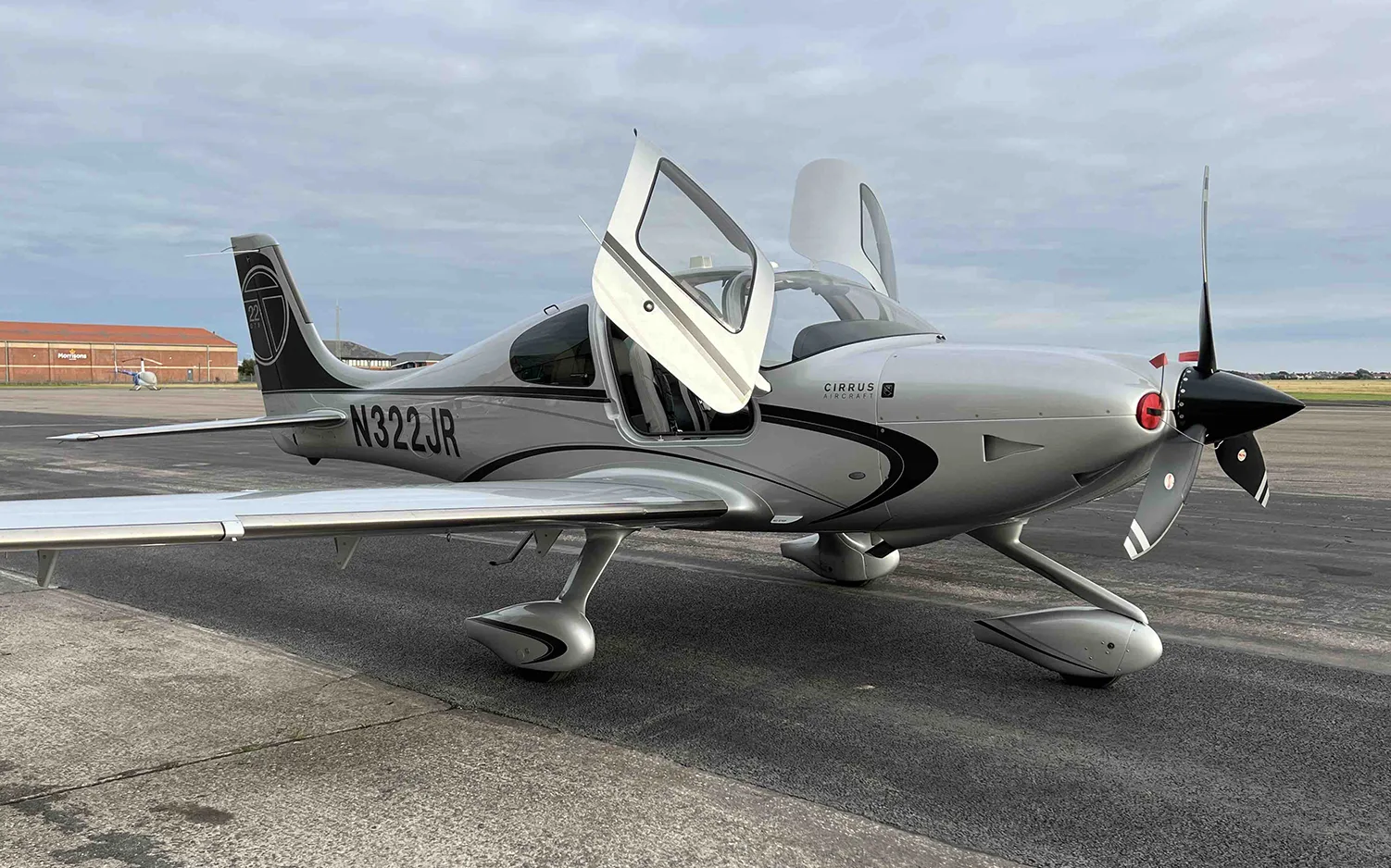 A silver Cirrus SR22 aircraft with registration N322JR and open doors is parked on an airfield.