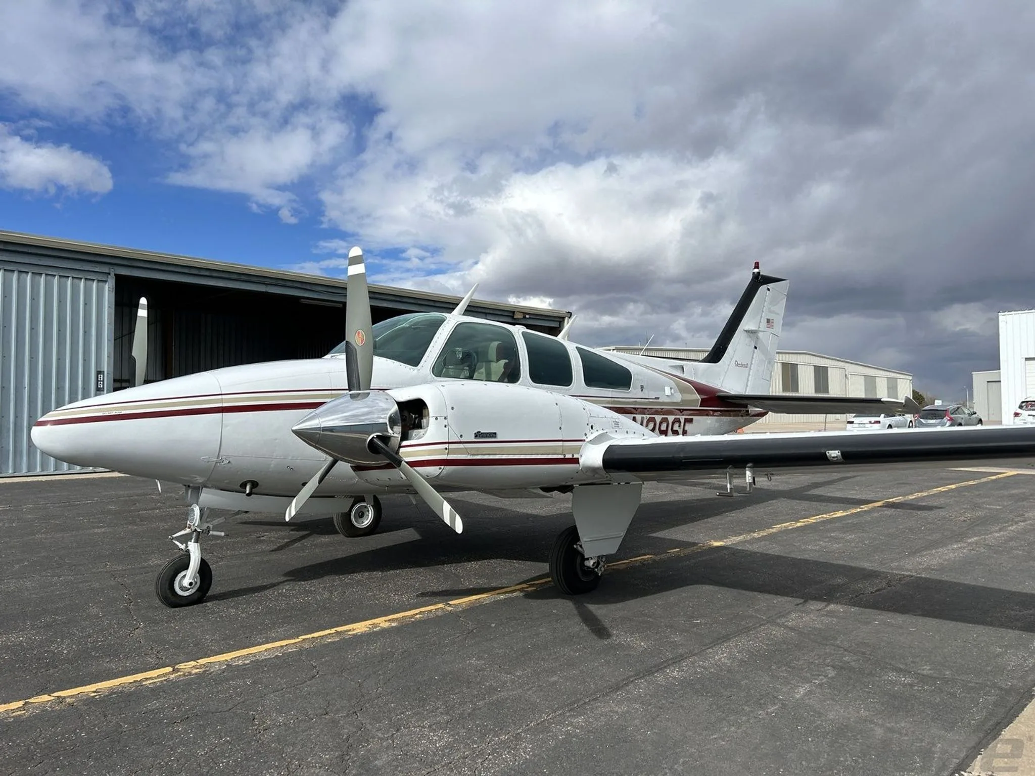 A white and maroon Beechcraft Baron N29SF parked on an asphalt ramp, viewed from the front left, with hangars in the background.