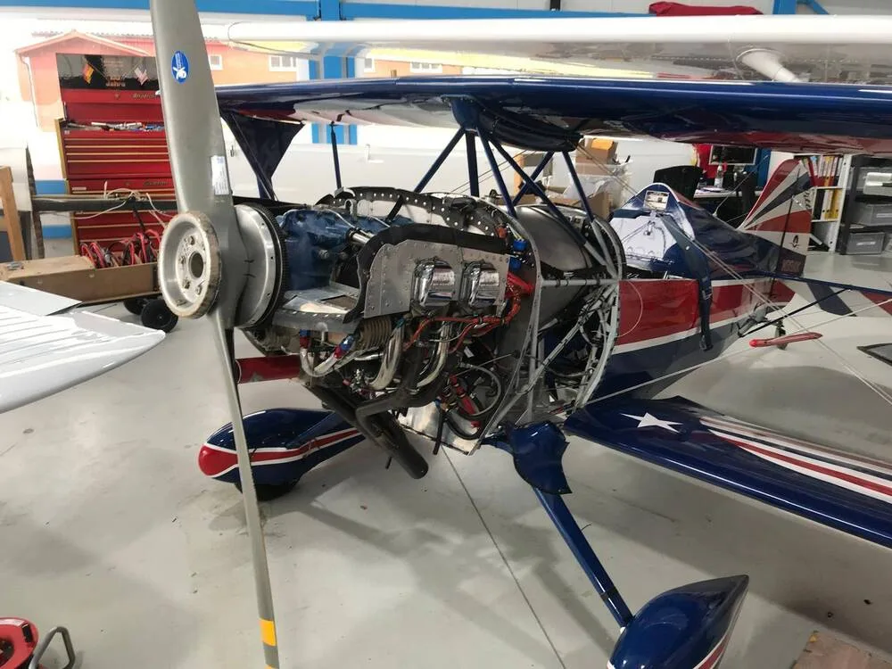 The engine of a red, white, and blue biplane, N99AK, is exposed for maintenance in a hangar.