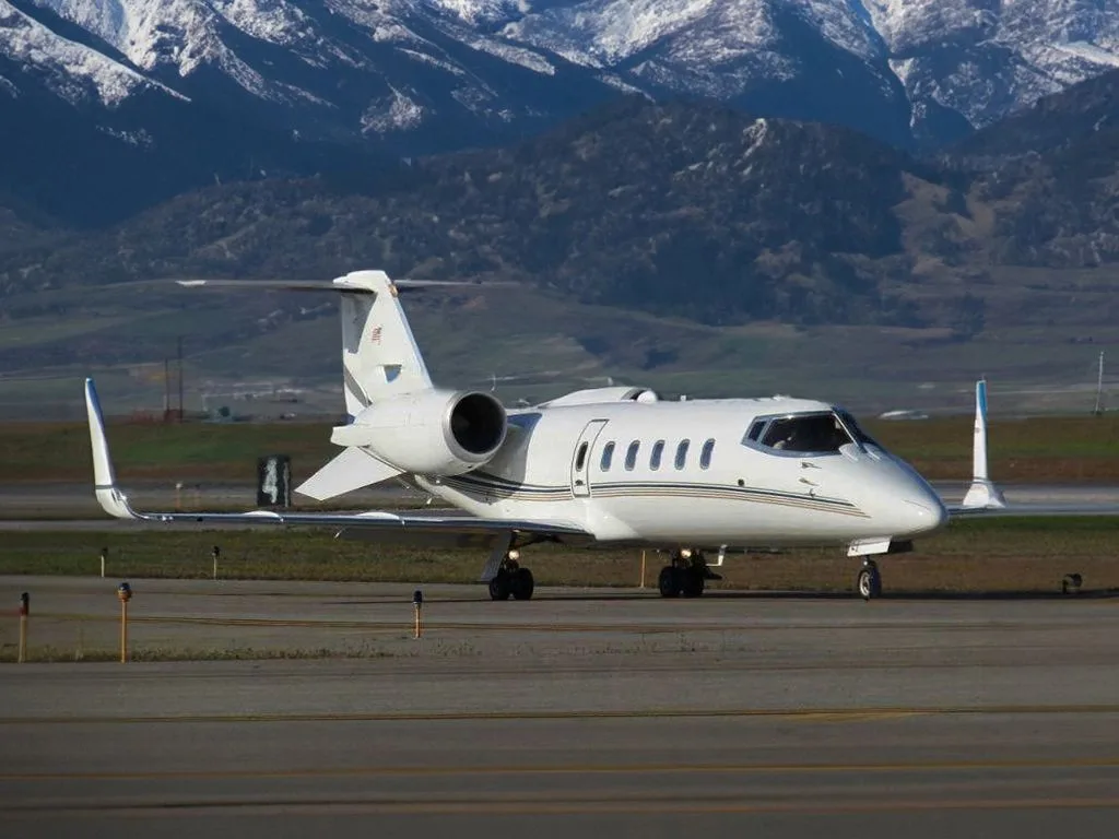A white Learjet business jet with a dark stripe on the fuselage, taxiing on an airfield with snow-capped mountains in the background.