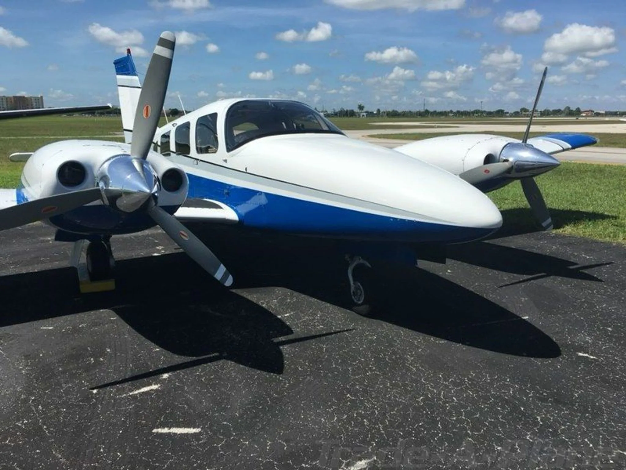 A white and blue twin-engine aircraft with propellers parked on an asphalt tarmac under a partly cloudy sky.
