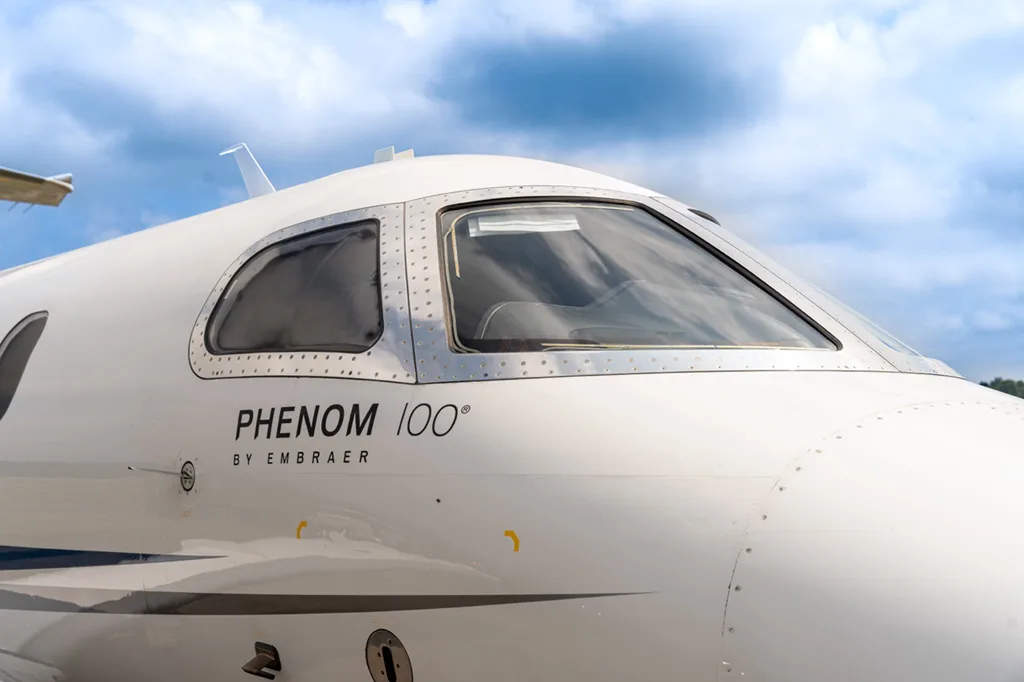 A close-up of the cockpit windows and nose of an Embraer Phenom 100, showing the aircraft model name.