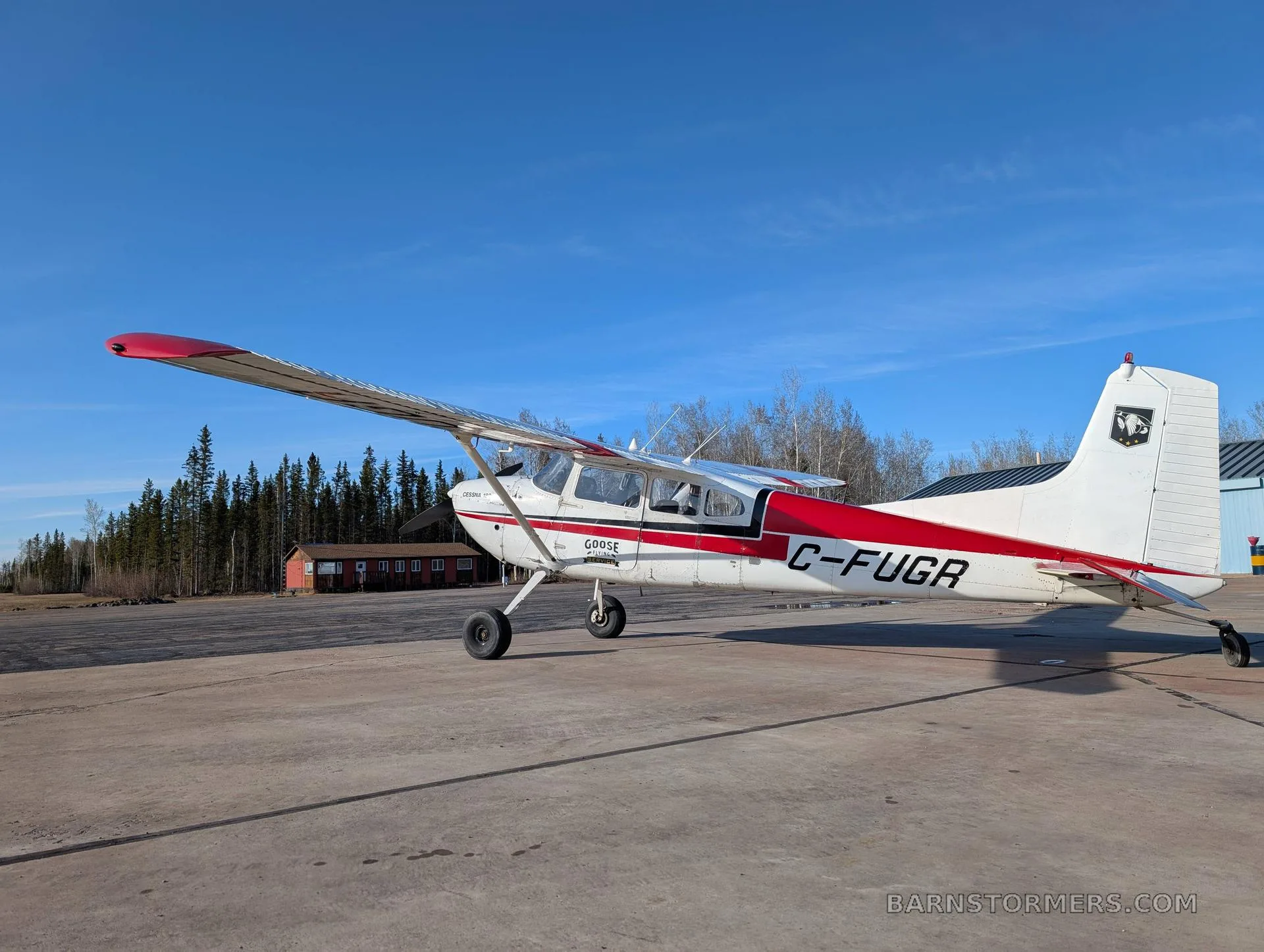 A white and red Cessna 180 (C-FUGR) is parked on a concrete tarmac with a red building and trees in the background.