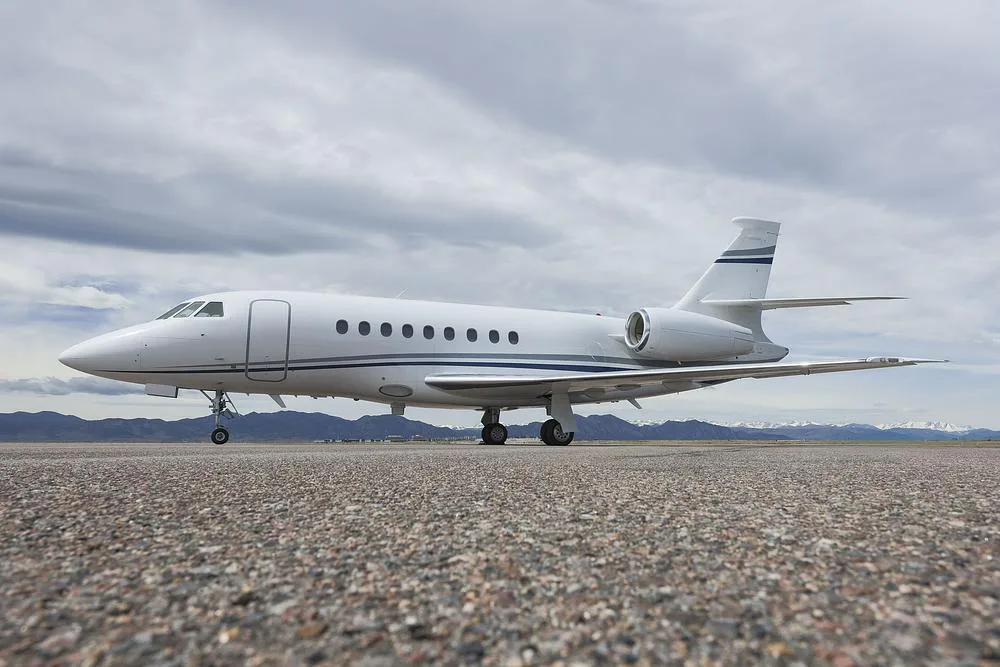 A white Dassault Falcon 900 business jet with blue and grey stripes parked on an asphalt runway under a cloudy sky, with mountains in the background.