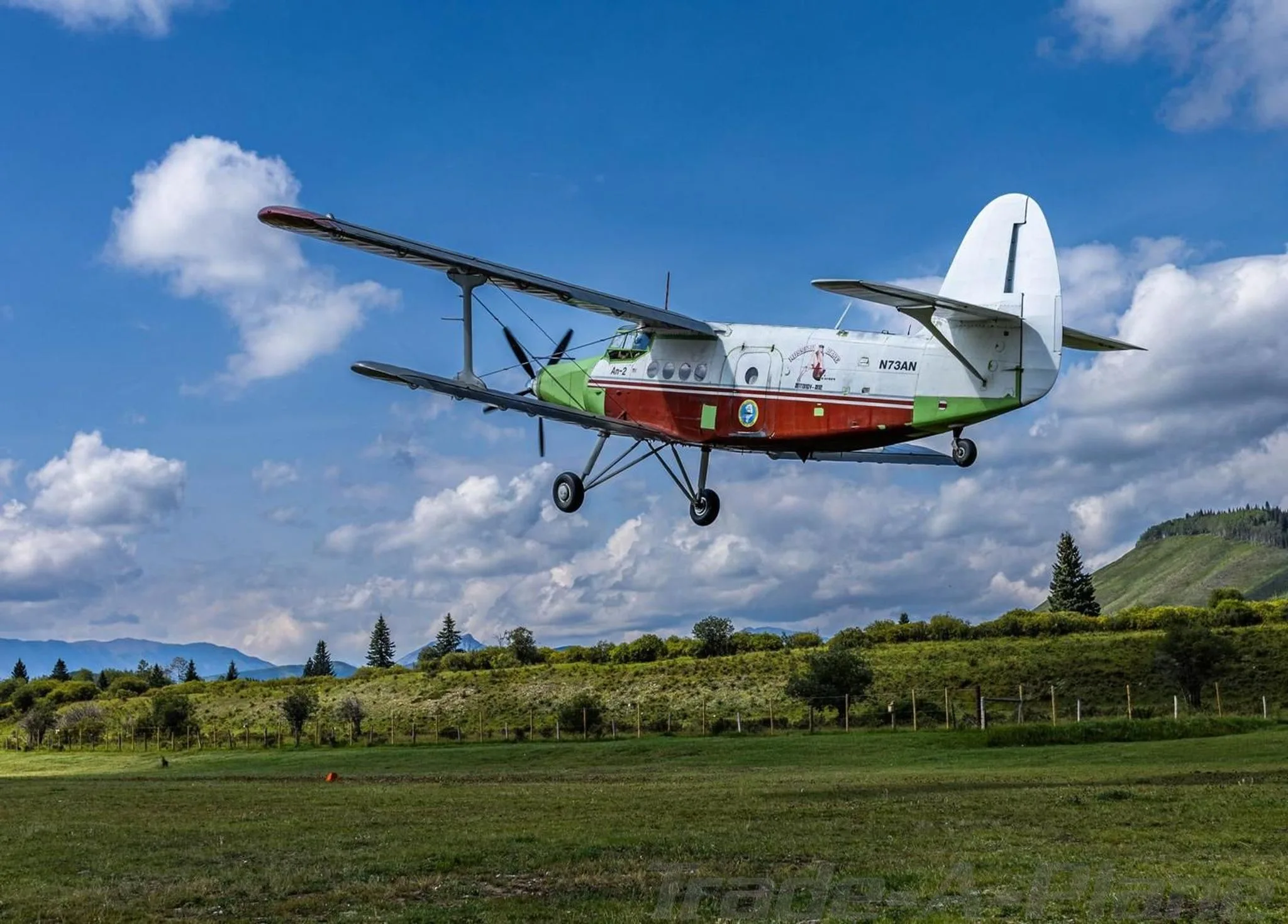 An Antonov An-2 biplane, painted white, red, and green, flying low over a green field with mountains in the background.