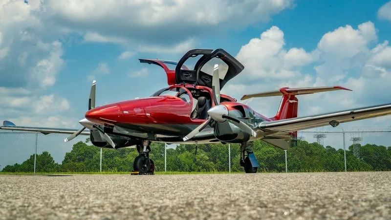 A red and grey Diamond DA62 twin-engine aircraft with its gull-wing doors open, parked on an airfield under a partly cloudy sky.