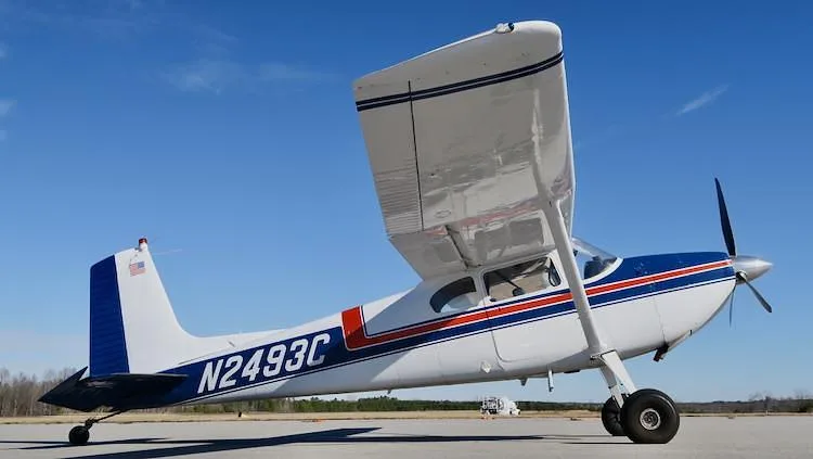 A three-quarter rear view of a white and blue Cessna 180 Skywagon with red stripes, registration N2493C, parked on an airfield.