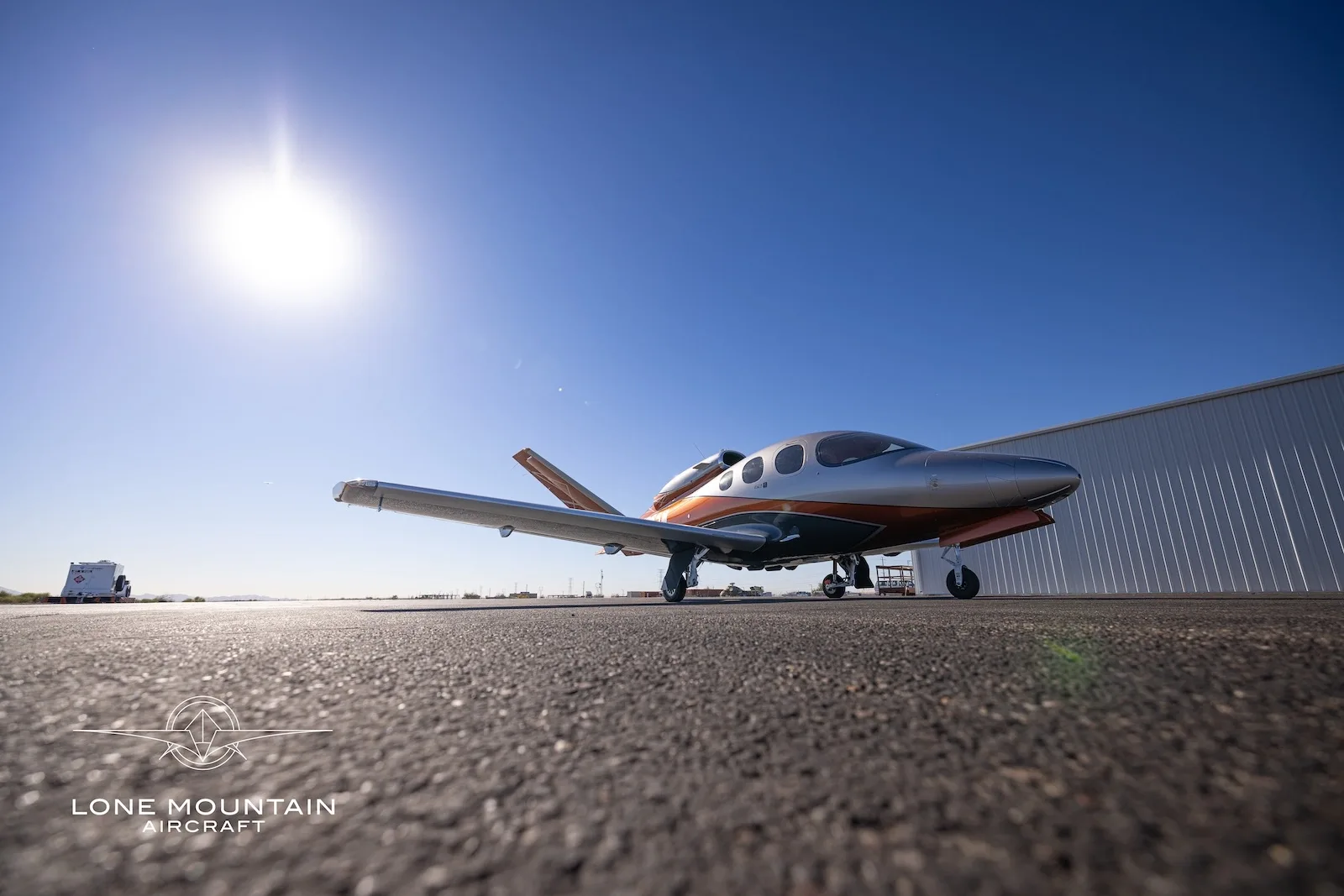 A low-angle view of a silver and orange Cirrus Vision Jet (SF50) parked on an asphalt tarmac under a bright sky.