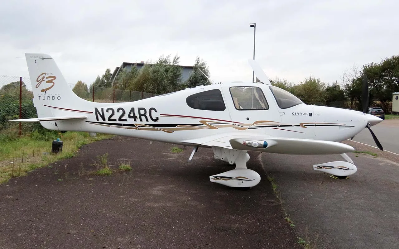 A white Cirrus SR22T (N224RC) with brown and black stripes parked on an asphalt surface next to a grassy area.