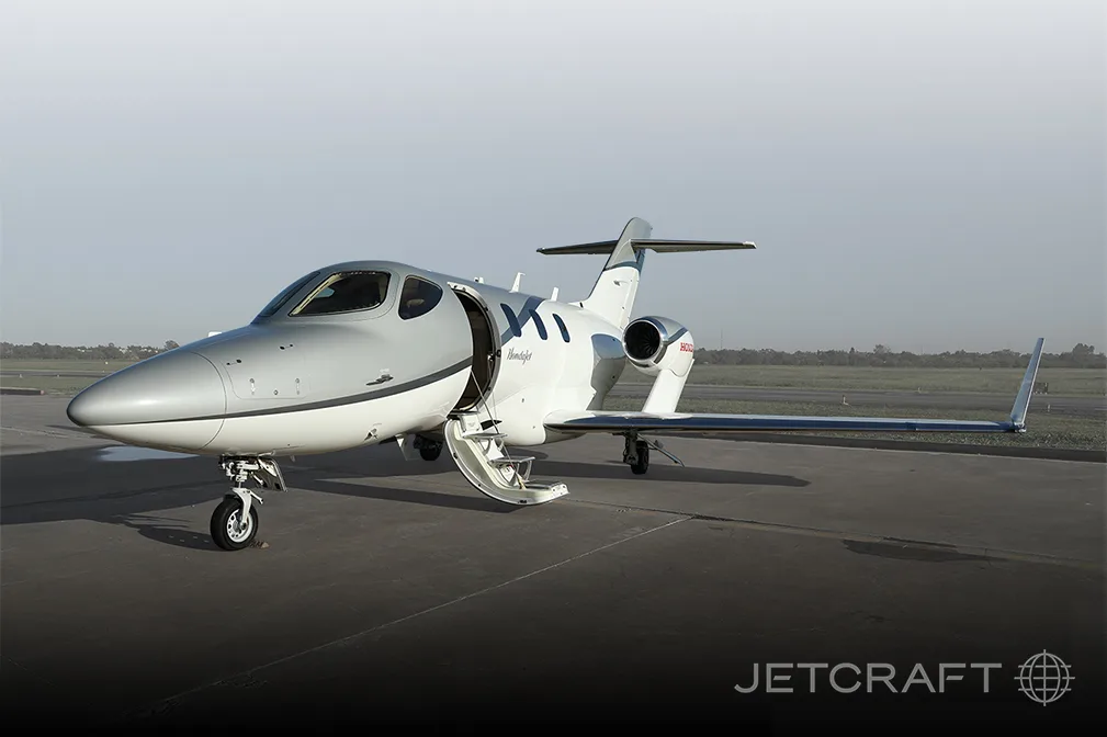 A silver and white HondaJet HA-420 is parked on an airfield with its main cabin door open.