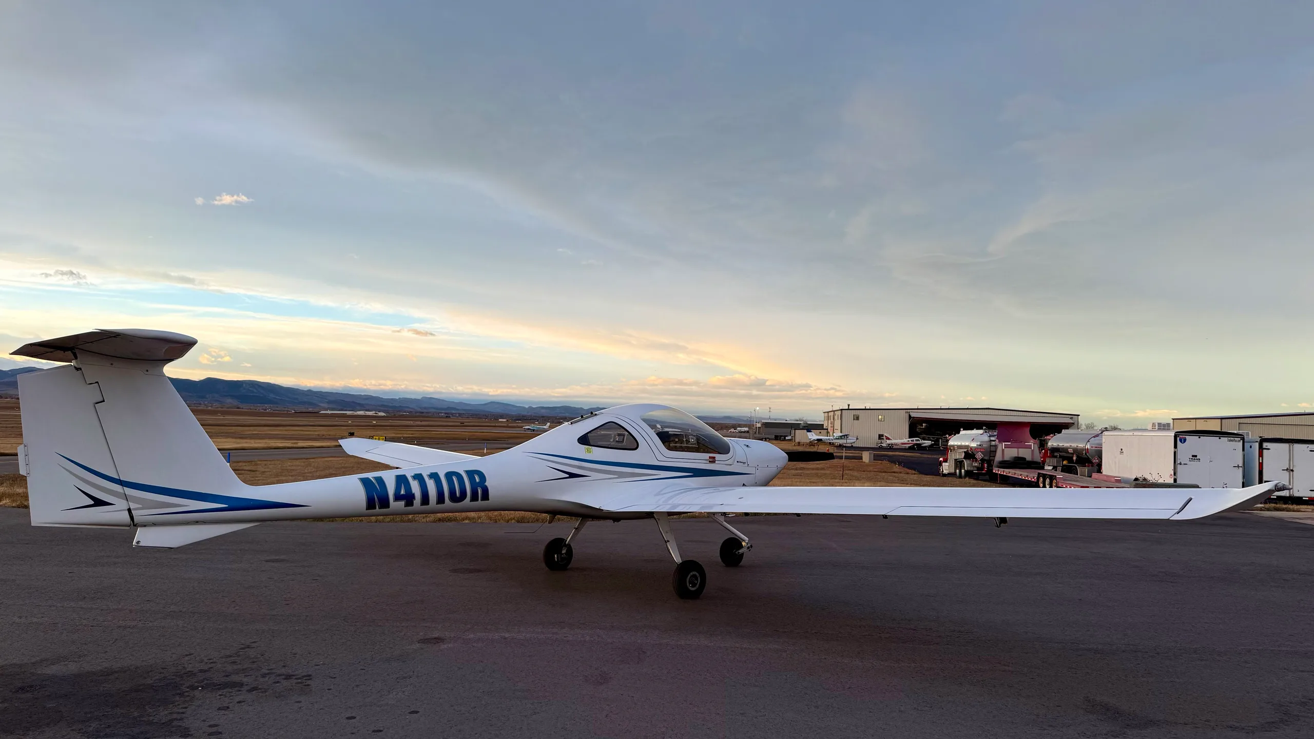 A white Diamond DA20-C1 Eclipse aircraft with blue stripes, registration N4110R, parked on an airfield with other aircraft and hangars in the distance.