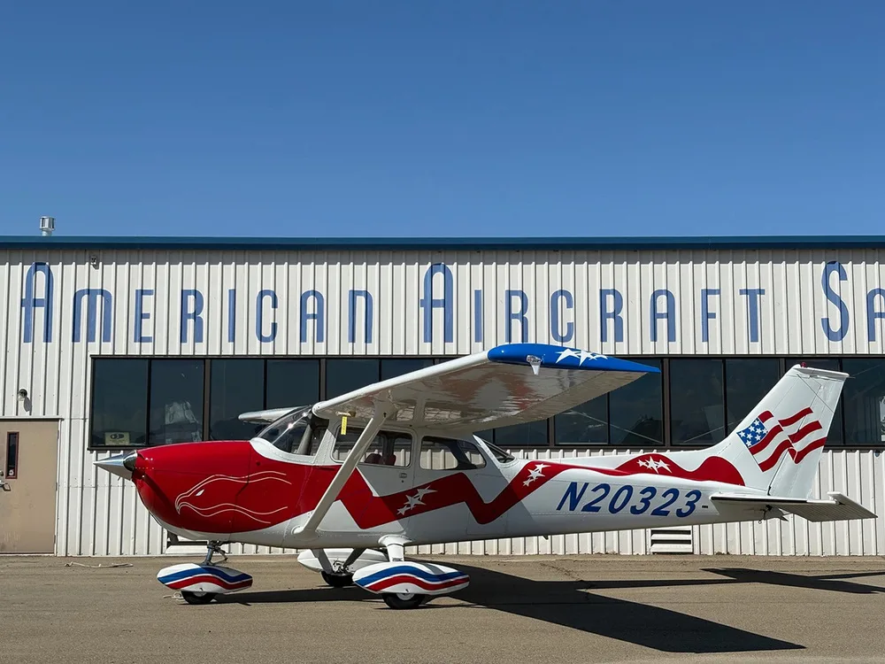 A side view of the red, white, and blue Cessna 172, N20323, parked on an airfield in front of a hangar.