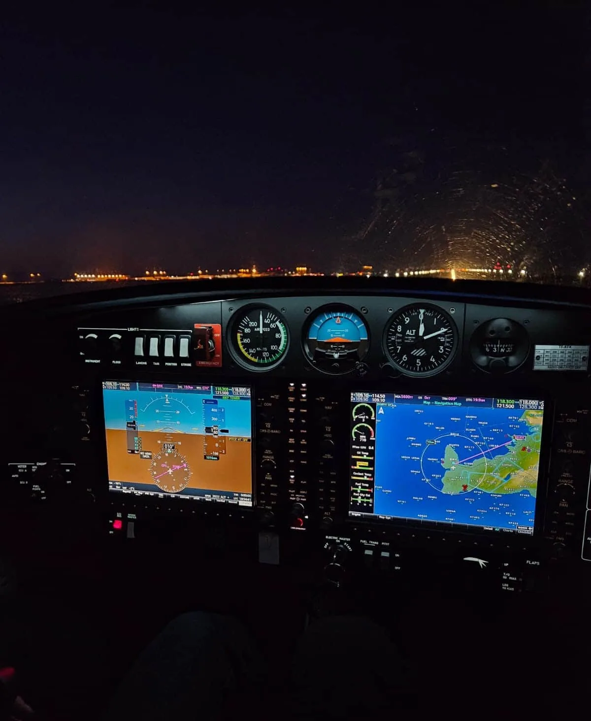 A night view from an aircraft cockpit, showing the illuminated instrument panel with multiple screens and the distant runway lights through the windshield.