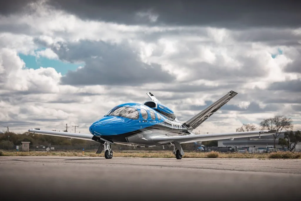 A blue and silver Cirrus Vision Jet parked on an airfield under a cloudy sky.