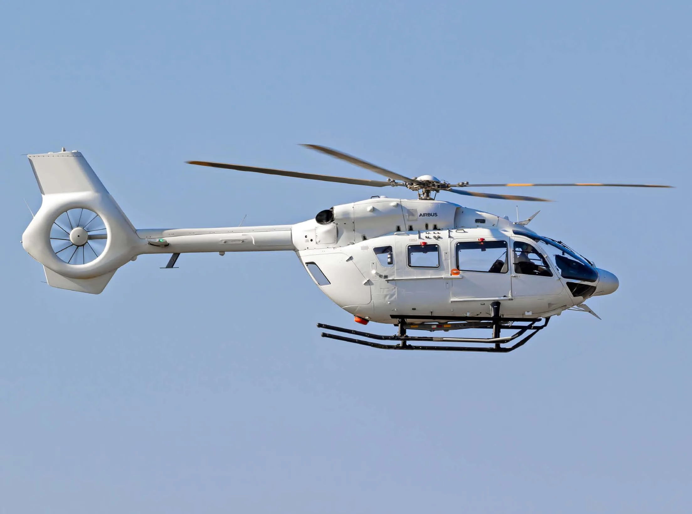 A white Airbus H135 helicopter with a Fenestron tail rotor is seen flying against a clear blue sky.